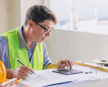 A man in glasses, blue shirt and yellow PPE vest sits working at a desk.