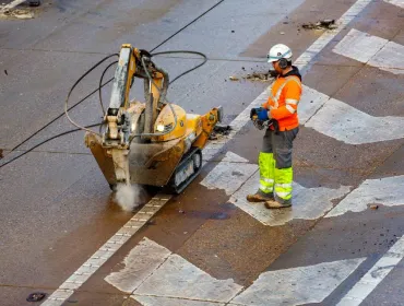 A man in orange, grey and yellow PPE uses a remote controlled piece of construction equipment fitted with a breaker to carry out repairs to a concrete road