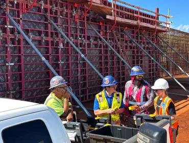 A group of construction workers in various colours of PPE stand in front of a wall of formwork
