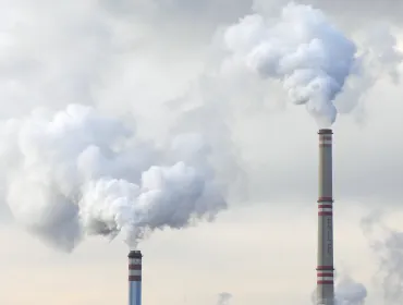 The top of two tall chimneys against a grey sky billow "smoke" into the atmosphere