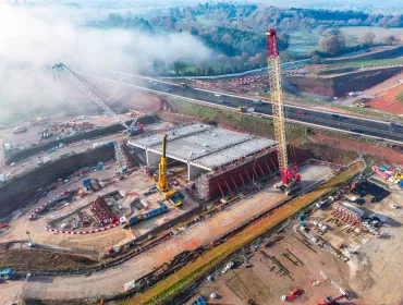 An aerial view of the A46 Kenilworth Bypass box structure under construction with a large yellow and red crawler crane and a smaller white telescopic crane and white crawler crane. Image: HS2.