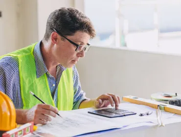 A man in glasses, blue shirt and yellow PPE vest sits working at a desk.