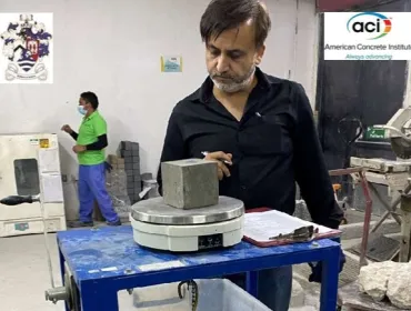 A man with a beard, waring a dark shirt inspects a concrete cube in a testing laboratory.