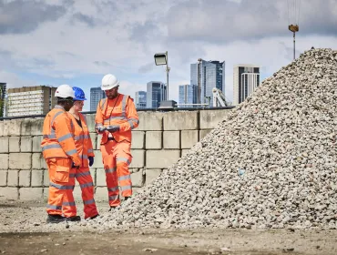 Three people in orange PPE stand discussing aggregate needs in a construction materials yard with a wall and city scape in the background