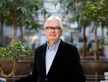 A grey-haired Sir Terry Farrell (1938-2025) wearing glasses, a dark jacket and light blue shirt photographed in front of an avenue of small trees in an urban setting © richard gleed / farrell