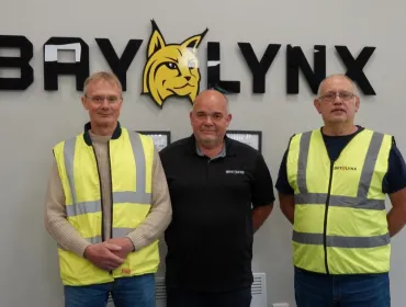 Three men stand in front of the BayLynx logo wearing PPE