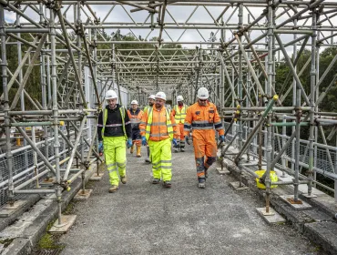 A group of construction workers walk along a concrete walkway, through a "tunnel" of scaffolding, wearing a variety of PPE , including orange hi vis jackets, yellow trousers, white hard hats and eye protectors