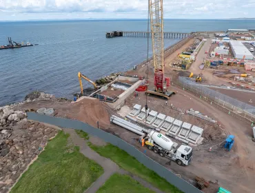 A red and yellow crawler crane lifts precast concrete elements at a shoreline construction site
