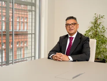 Vijay Bange in a dark suit, white shirt and red tie sits at an empty table with windows behind him showing a red brick building.
