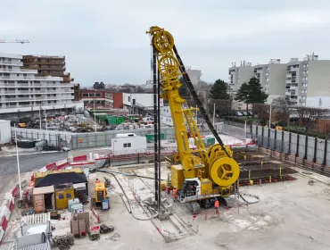 An elevated view of a yellow Soilmec SC-110 Urban cutter working on the Grand Paris Express, constructing 1,200mm-thick diaphragm walls up to 38 metres deep at Rueil–Suresnes Mont-Valérien station, surrounded by houses, low rise buildings and roads.