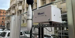 a dust monitoring station attached to a metal fence withan building in the background