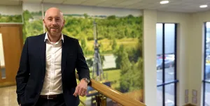A bald man with a beard in an open necked shirt and dark suit standing on an internal office landing