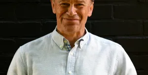 A head and shoulders picture of a man with unruly hair in a white shirt against a black background