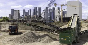 Piles of aggregate in a yard with a truck and green screen with a crane and tall buildings in the background