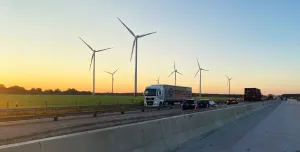 A truck on a motorway with a concrete safety barrier in the foreground and several wind turbines in the background.