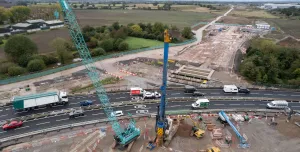 A green crane and yellow piling rig photographed from the air installing piling on a large construction site by a dual carriageway