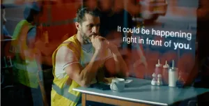 A man with dark hair in a top knot, wearing yellow PPE sitting at a cafe table looking anxious