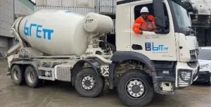 A white concrete mixer truck being driven by a man in red PPE
