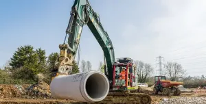 A green excavator with yellow pipe-handling grab moves a large piece of concrete pipe