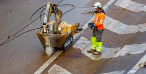 A man in orange, grey and yellow PPE uses a remote controlled piece of construction equipment fitted with a breaker to carry out repairs to a concrete road