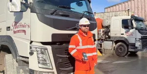 A man in orange PPE standing in front of a cement mixer holding a mobile device that uses the Podfather app to access their schedules and delivery notes, navigate routes, and record delivery details digitally. 