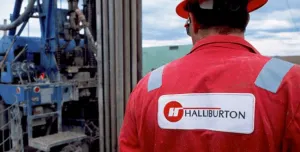 A man in red overalls and red hard hat stands with his back to the camera, a Halliburton logo on his back, surveying a Halliburton Energy Services project.