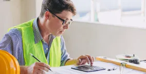 A man in glasses, blue shirt and yellow PPE vest sits working at a desk.