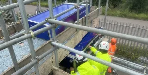 Two people in yellow PPE carry out a bridge inspection from a scaffold suspended from the side of the bridge