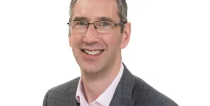A head and shoulders shot of Peter Reekie of the Scottish Futures Trust will speak on sustainable infrastructure at the Glasgow Industry Day wearing glasses, an open necked pick shirt and a grey over-check jacket