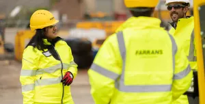 Three people in yellow PPE stand talking with construction machinery in the background