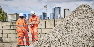Three people in orange PPE stand discussing aggregate needs in a construction materials yard with a wall and city scape in the background