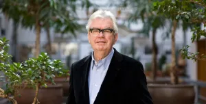 A grey-haired Sir Terry Farrell (1938-2025) wearing glasses, a dark jacket and light blue shirt photographed in front of an avenue of small trees in an urban setting © richard gleed / farrell