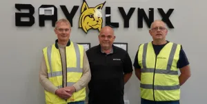 Three men stand in front of the BayLynx logo wearing PPE
