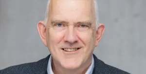 Head and shoulders photo of a bald, smiling man in open necked shirt, jumper and jacket