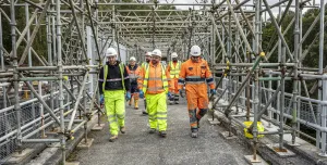 A group of construction workers walk along a concrete walkway, through a "tunnel" of scaffolding, wearing a variety of PPE , including orange hi vis jackets, yellow trousers, white hard hats and eye protectors