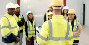 A group of youg people wearing yellow PPE and white and yellow hard hats listen to an older worker