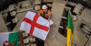 A man wearing a hard hat and waving a Cross of St George flag pokes his head through a TBM cutter wheel with a Welsh flag and another unidentified flag adorning the cutter head as HS2 engineers celebrate the breakthrough of tunnel boring machine Elizabeth at Washwood Heath in Birmingham, completing the 3.5-mile Bromford tunnel under the M6. (Image © HS2 Ltd)