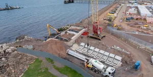 A red and yellow crawler crane lifts precast concrete elements at a shoreline construction site