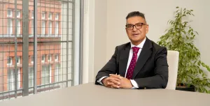Vijay Bange in a dark suit, white shirt and red tie sits at an empty table with windows behind him showing a red brick building.
