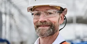 A head and shoulders photo of a man with a beard and glasses wearing PPE and a white hard hat