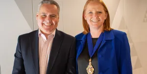 A smiling man and woman in business attire (dark suit, open necked shirt, blue jacket, black top) look at the camera