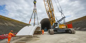 A yellow crawler crane lifts a precast concrete tunnel segment ready for placing on a cut and cover tunnel in the background
