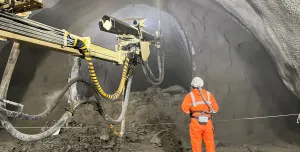 A construction worker in orange PPE uses a remote control unit to control a Normet concrete sprayer in a tunnel