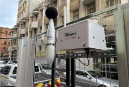 a dust monitoring station attached to a metal fence withan building in the background
