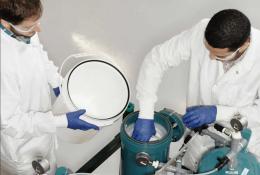 Two men in lab coats and safety glasses working in a lab next to two green canisters