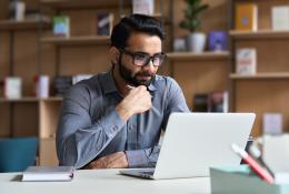 A man with a beard and glasses sitting at a desk looking at a laptop