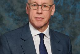 A studio head and shoulders photo of a man wearing glasses and a blue suit and tie