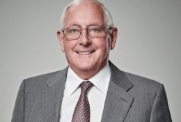 A head and shoulders picture of an old man in a grey suit and tie wearing glasses