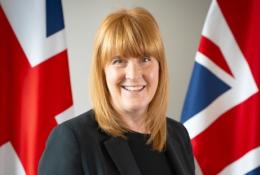 A head and shoulders photo of a woman with red hair and dark clothes standing in front of two Union Jack flags