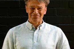 A head and shoulders picture of a man with unruly hair in a white shirt against a black background